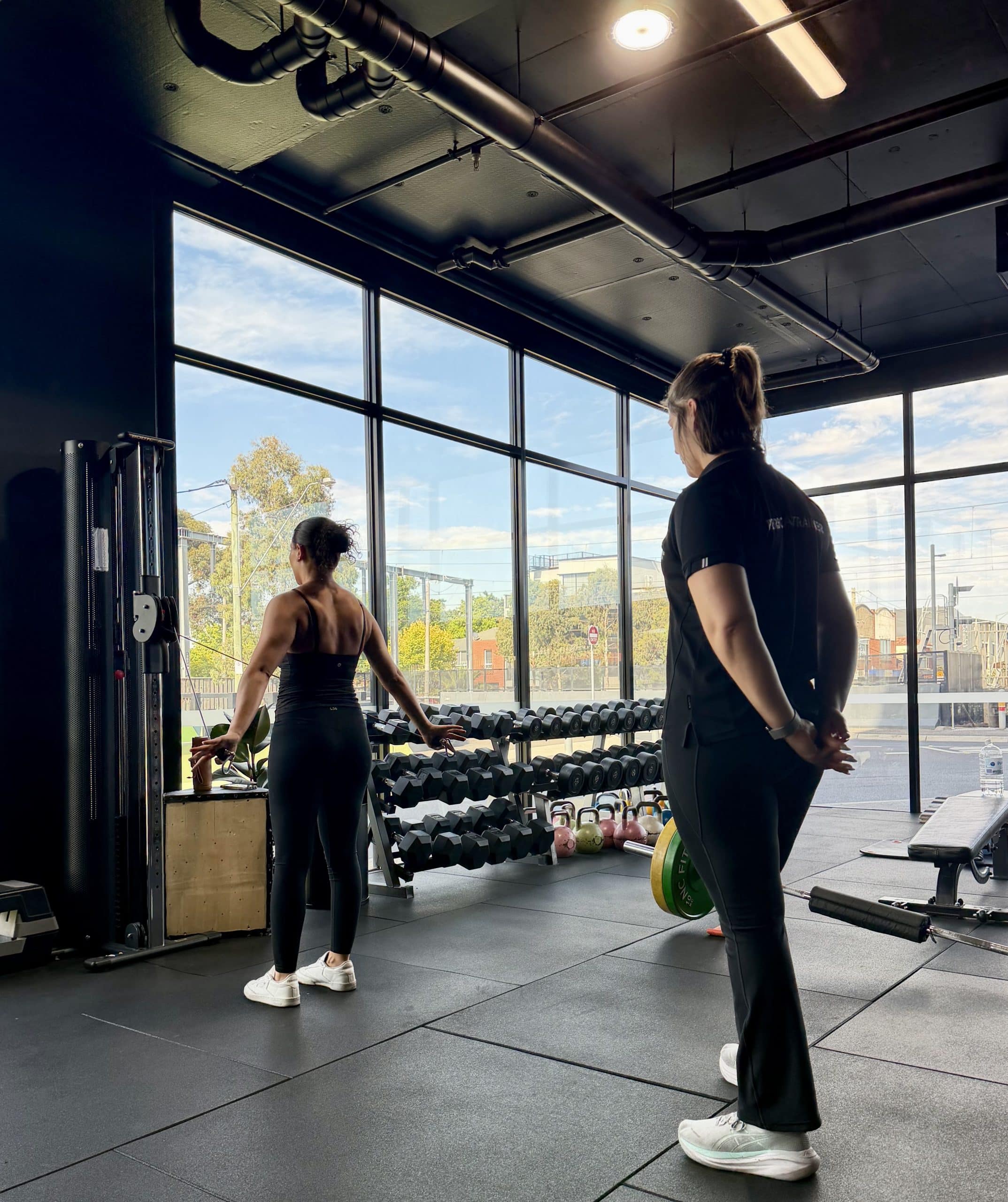 Female personal trainer supervising a client's cable exercise in a bright Essendon studio with large windows.
