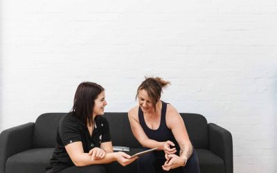 Trainer and client smiling on a couch while reviewing health and progress results on a tablet.